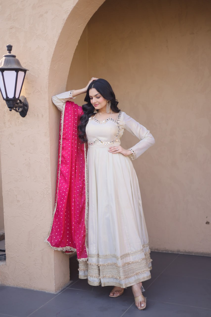 Woman in a white traditional outfit with a pink dupatta standing against a beige wall.