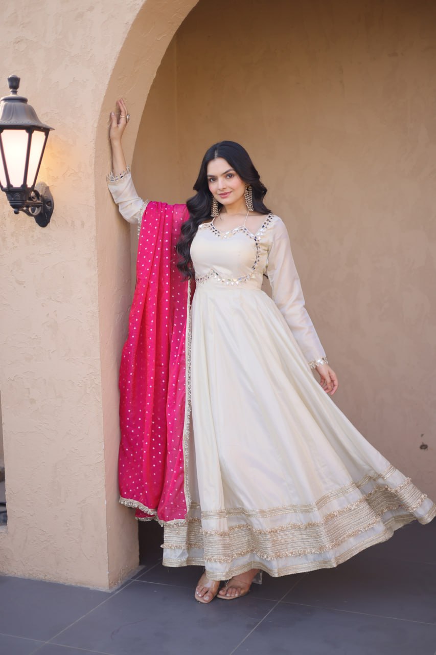 Woman in a white traditional outfit with a pink dupatta standing against a beige wall.