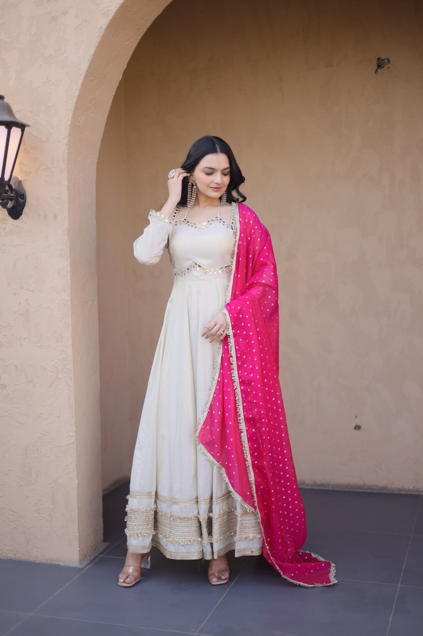 Woman in a white traditional outfit with a pink dupatta standing against a beige wall.