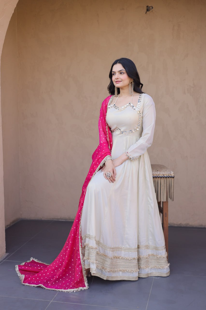 Woman in a white traditional outfit with a pink dupatta standing against a beige wall.