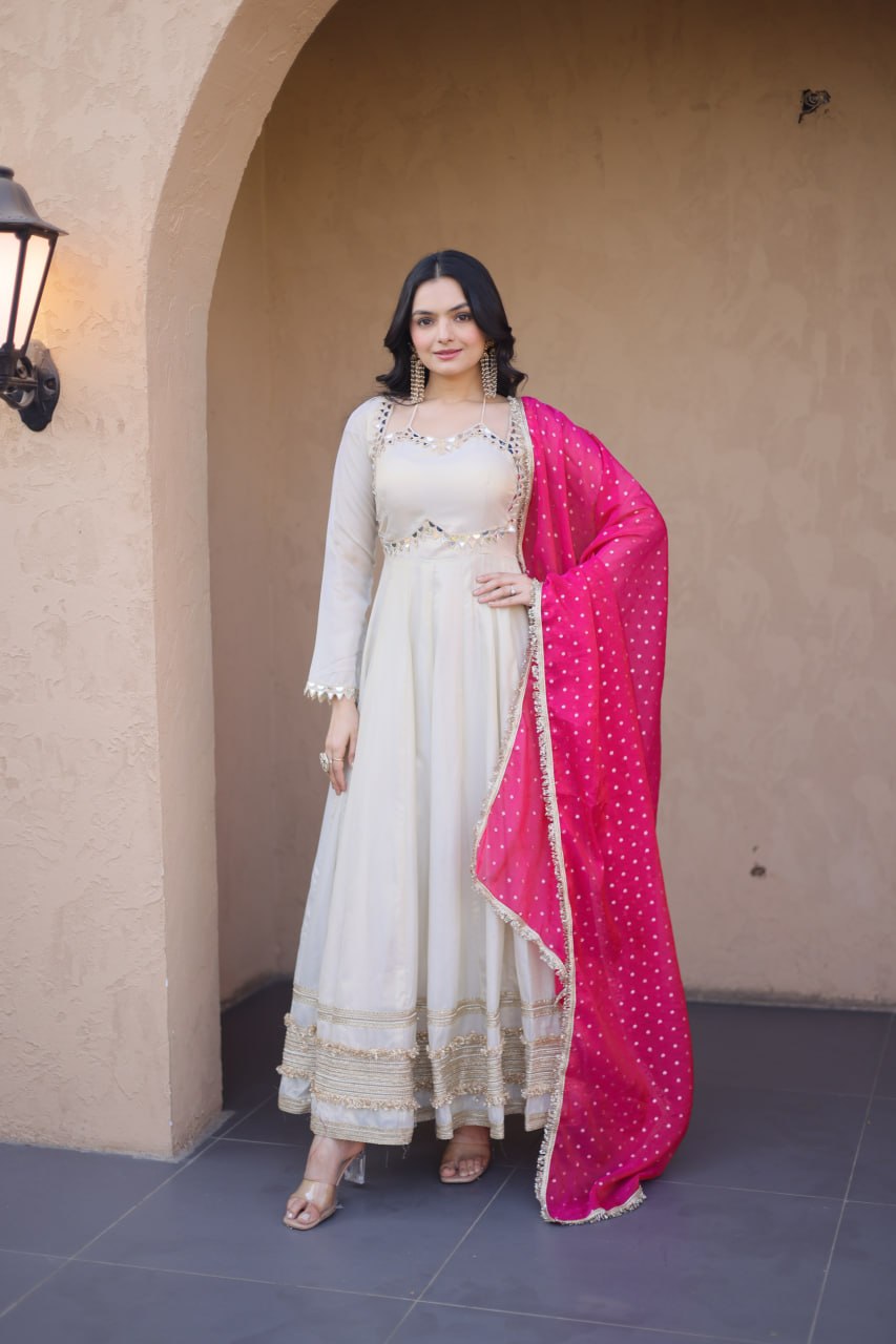Woman in a white traditional outfit with a pink dupatta standing against a beige wall.