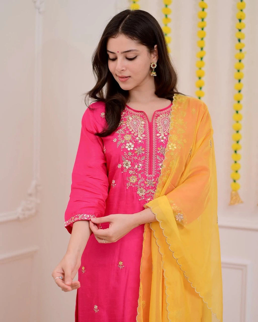 Woman wearing a pink embroidered outfit with a yellow dupatta against a white wall.