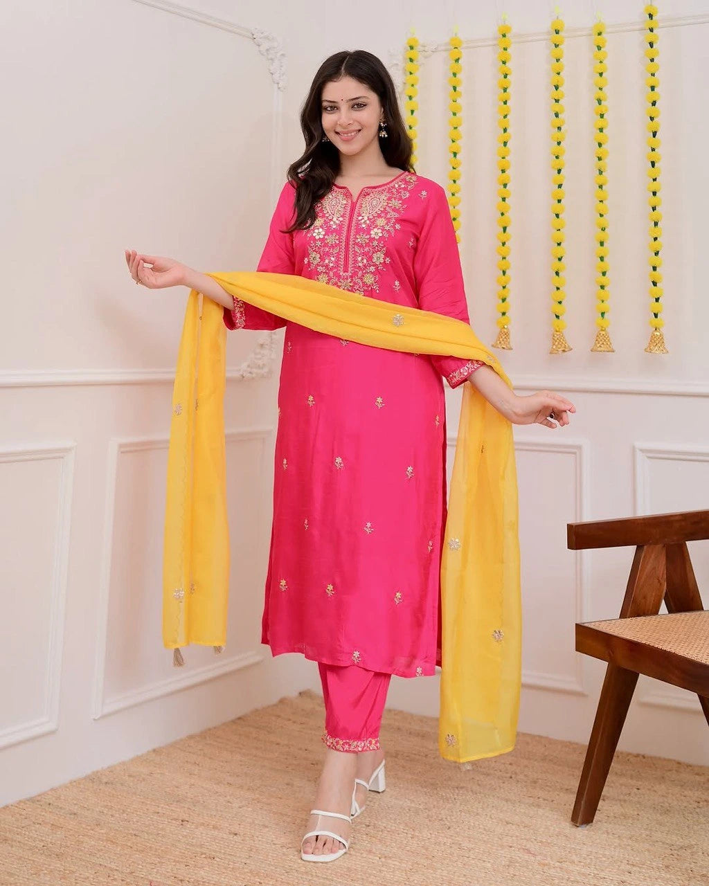 Woman in a pink traditional outfit with yellow dupatta in a room with decorative wall hanging.