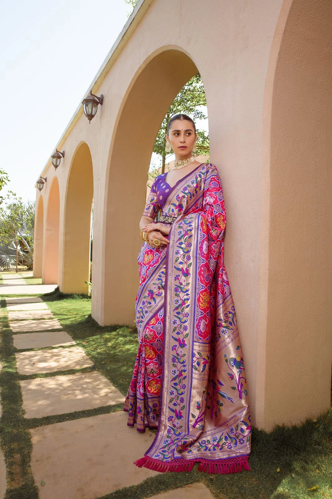 Woman in a colorful saree standing in front of a building with arches.
