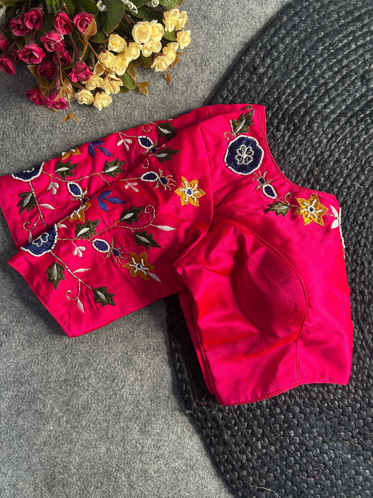 A front view of the pink silk blouse showcasing a deep U-shaped neckline. The neckline is bordered with dark blue and yellow embroidered flowers and detailed leaf patterns. The short sleeves are fully decorated with matching floral embroidery and gold-toned vines. The blouse is displayed on a textured grey surface next to a dark woven mat and a bouquet of small roses.