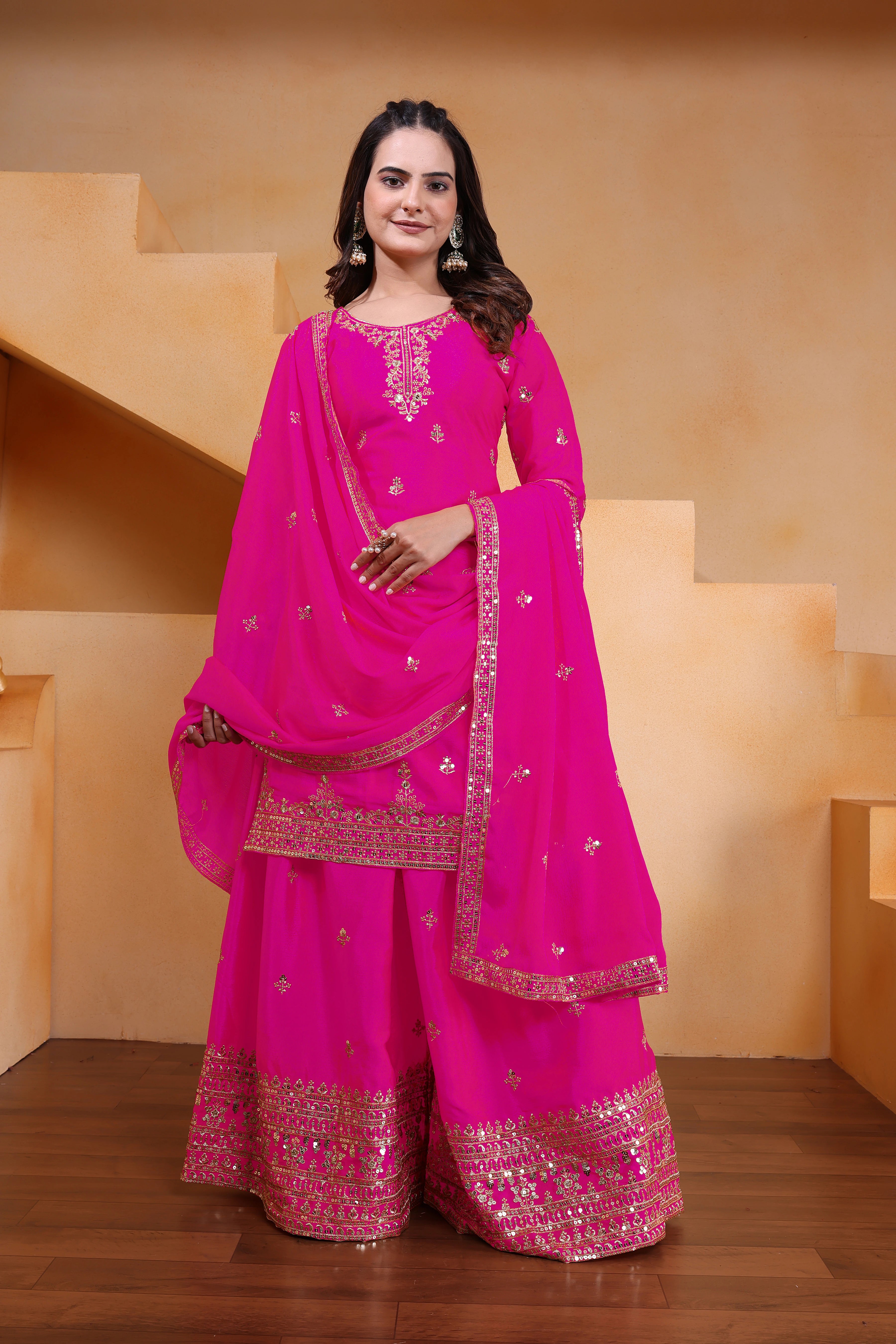 Woman in a bright pink traditional outfit with gold embroidery standing against a beige wall.