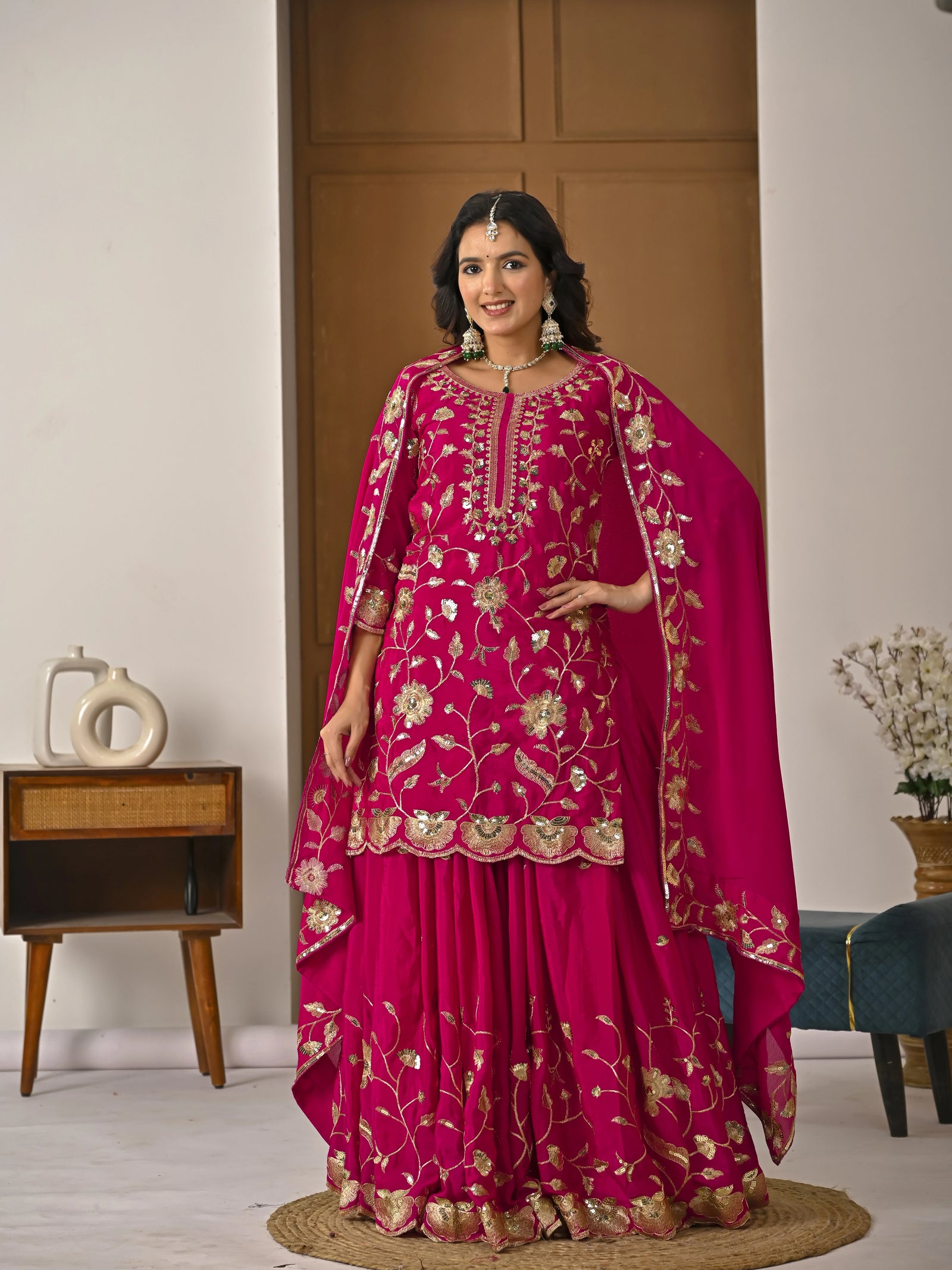 Woman in a pink embroidered traditional outfit standing in a room with wooden furniture and decor.