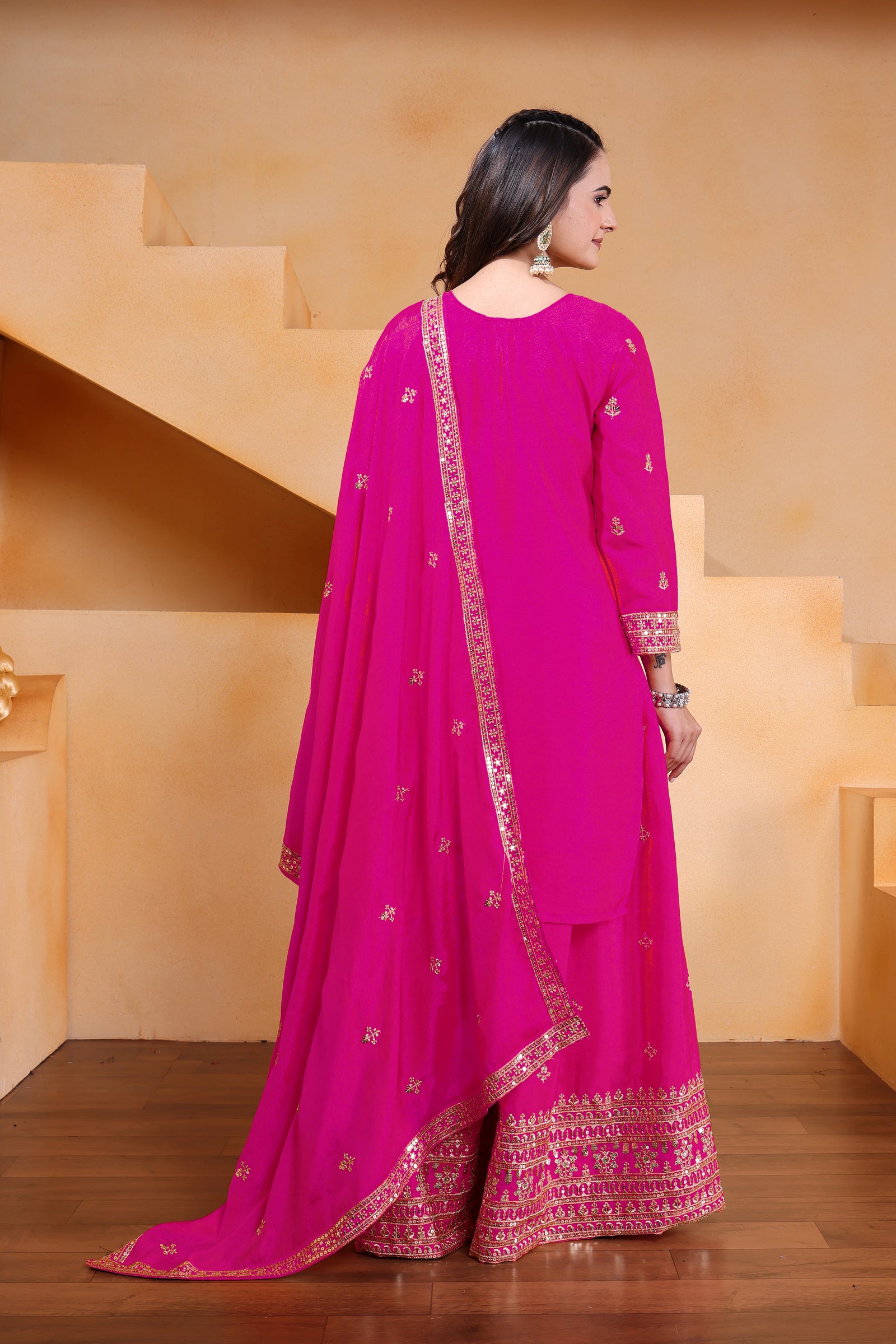 Woman wearing a bright pink traditional outfit with gold embroidery in a neutral setting.