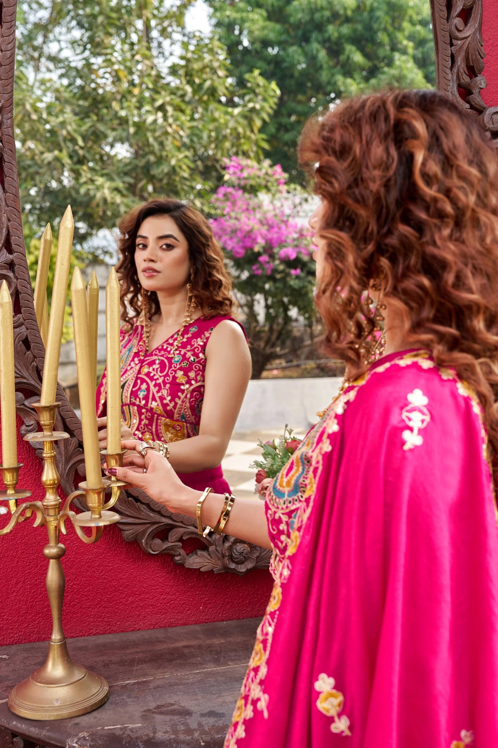 Woman in a pink traditional outfit adjusting candles in front of a mirror outdoors.