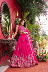 Woman in a pink traditional outfit standing in an outdoor setting with red walls and greenery.