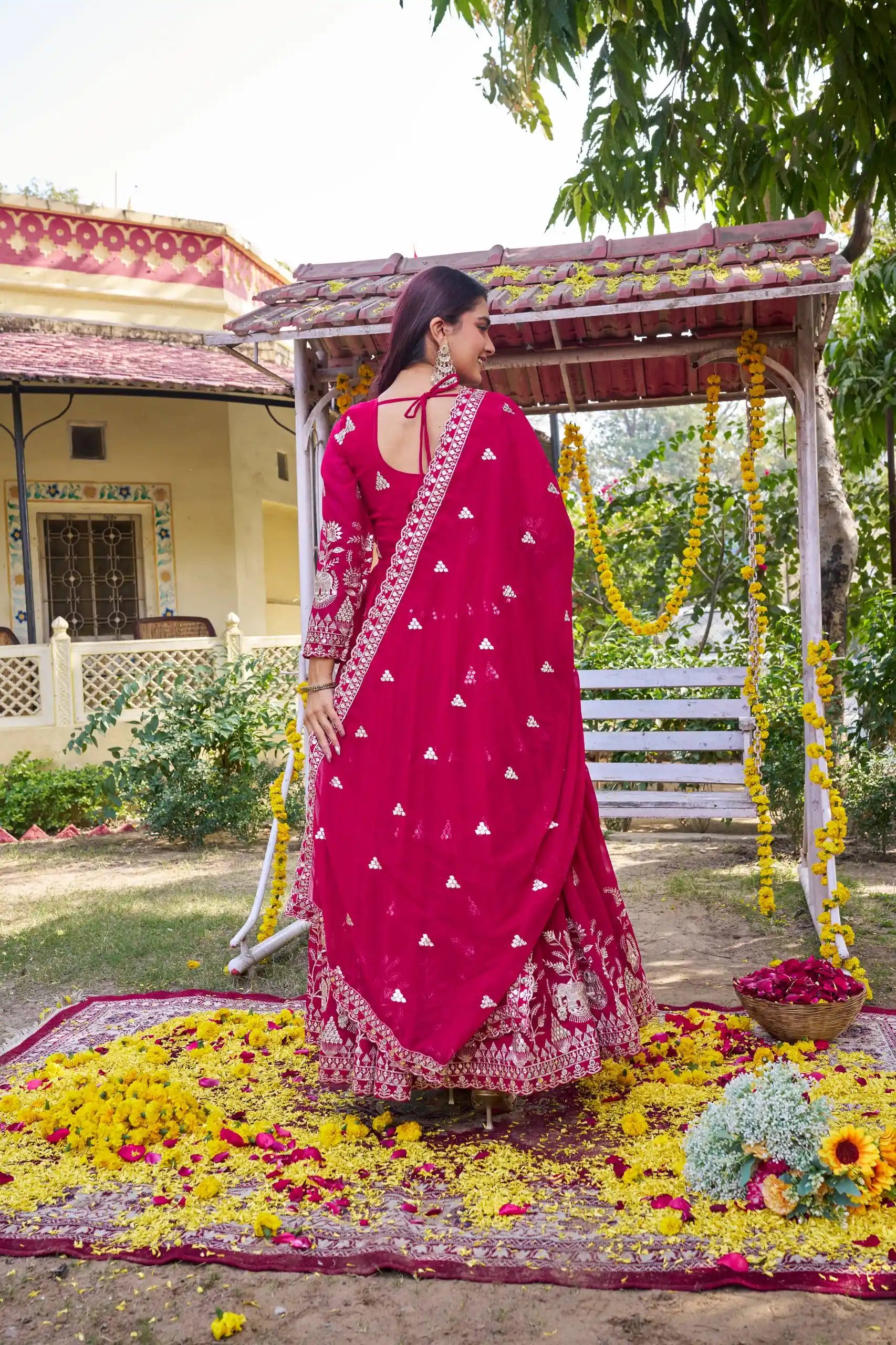 Woman in a pink saree standing under a decorated swing with flowers and a house in the background.