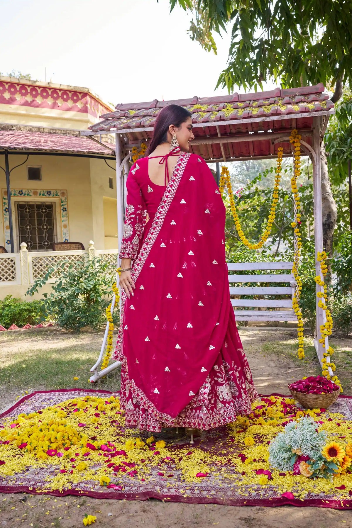 Woman in a pink saree standing under a decorated swing with flowers and a house in the background.