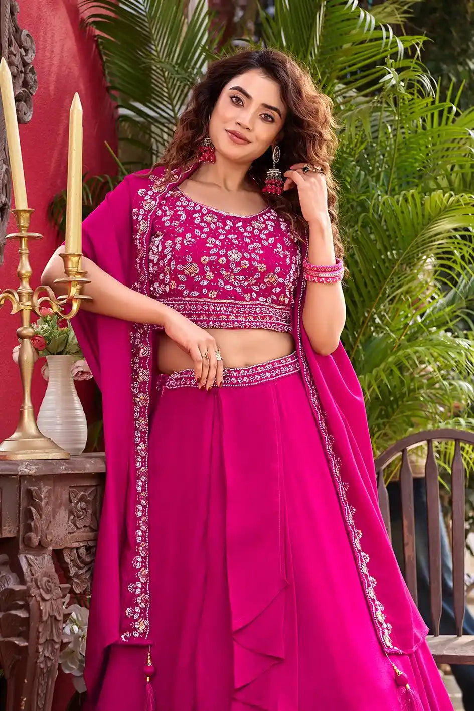 Woman in a pink traditional outfit with floral patterns, standing outdoors with greenery and a decorative table in the background.