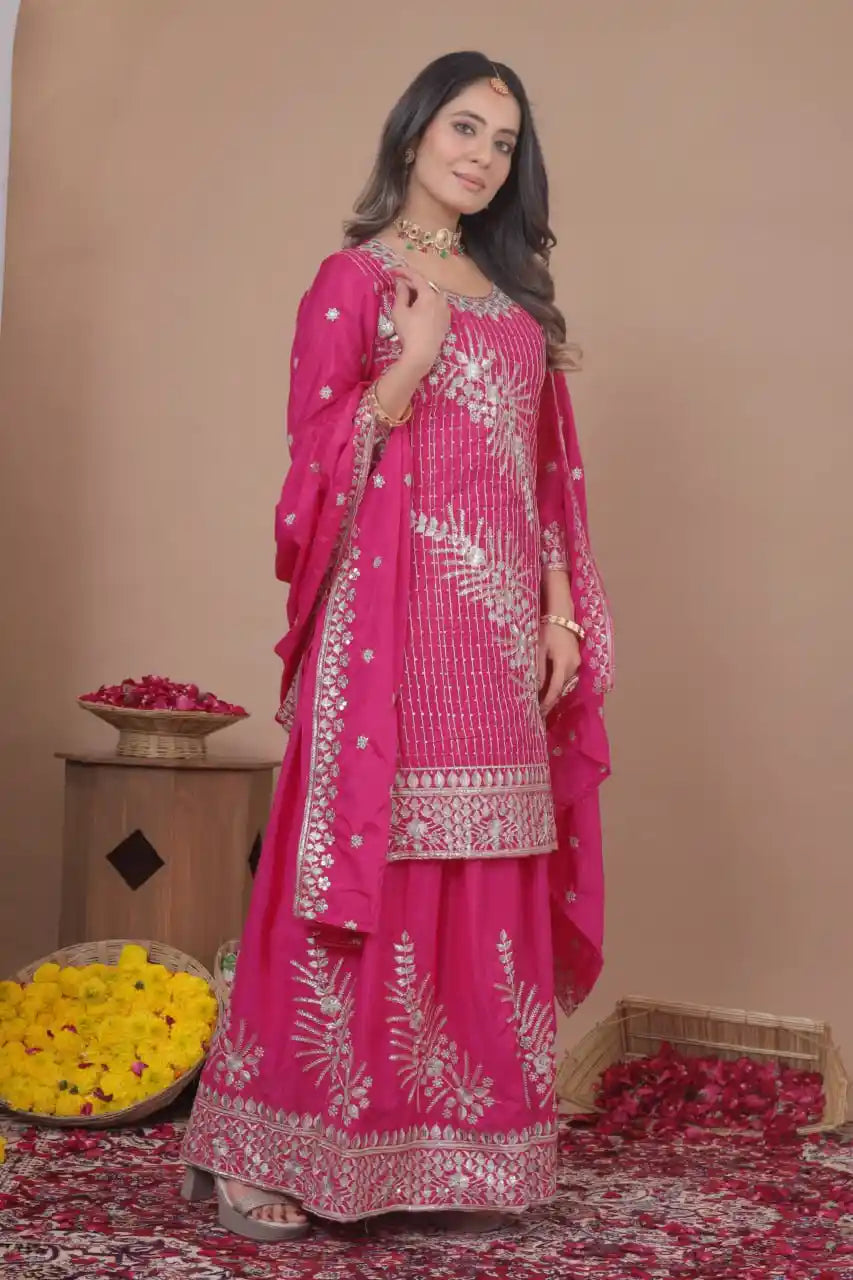 Woman in a pink traditional outfit with white embroidery standing against a beige wall.