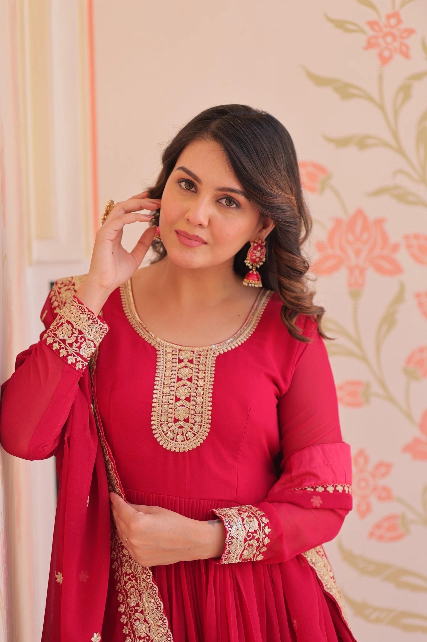 Woman wearing a red traditional outfit with gold embroidery against a floral wallpaper background