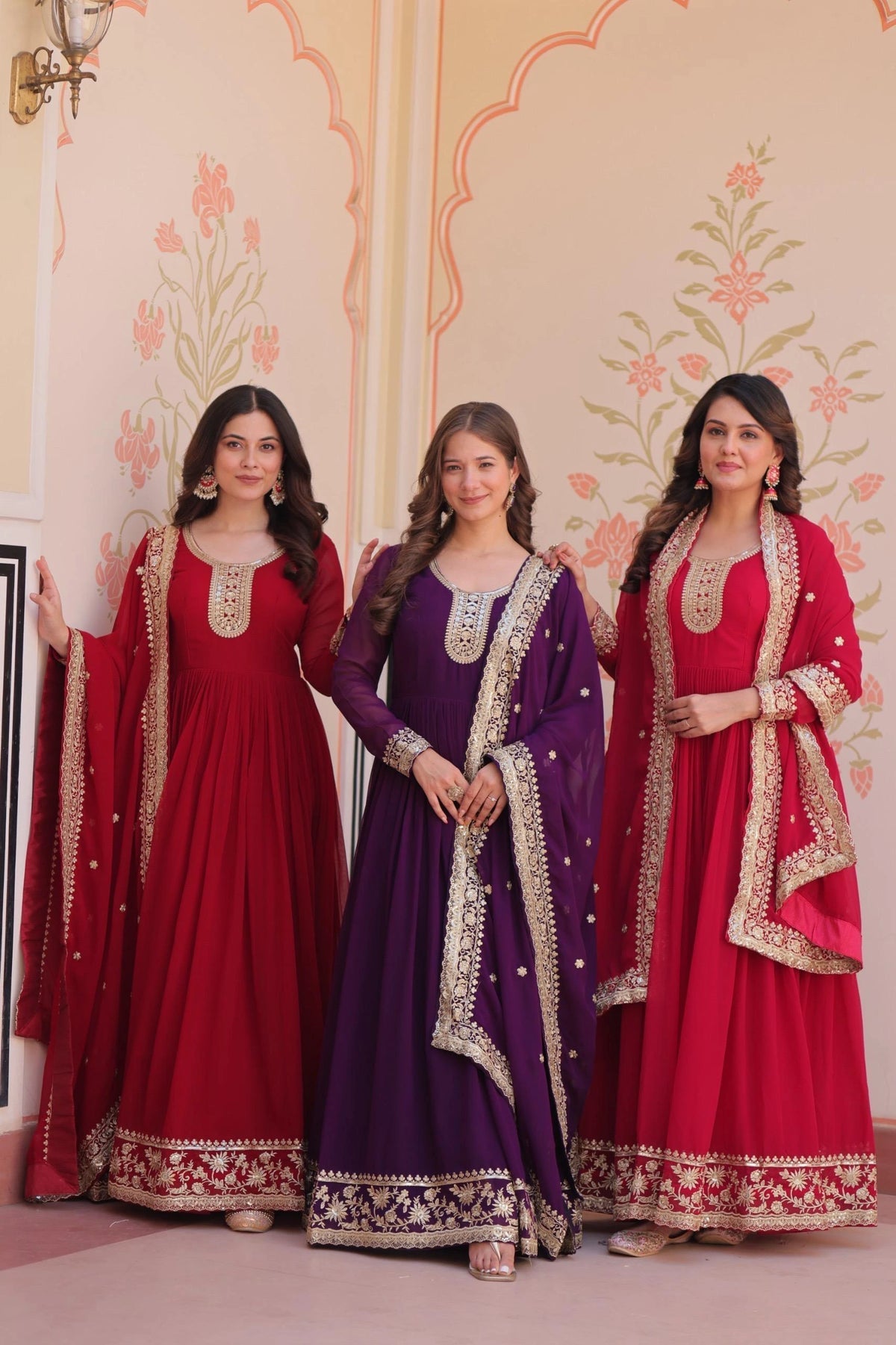 Three women in traditional red and purple dresses with gold embroidery standing against a decorative wall.