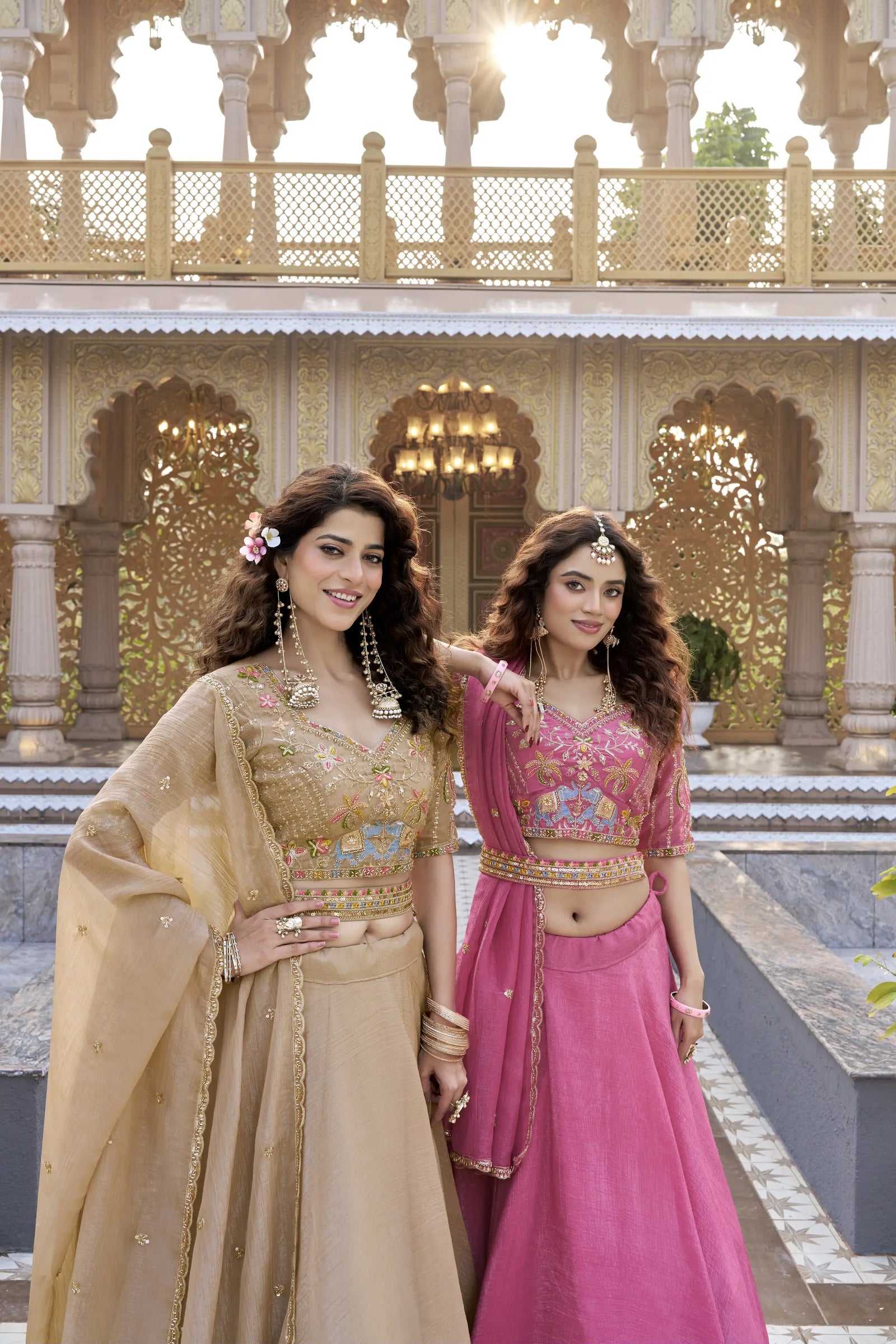 Two women in traditional Indian attire standing in front of an ornate architectural background.