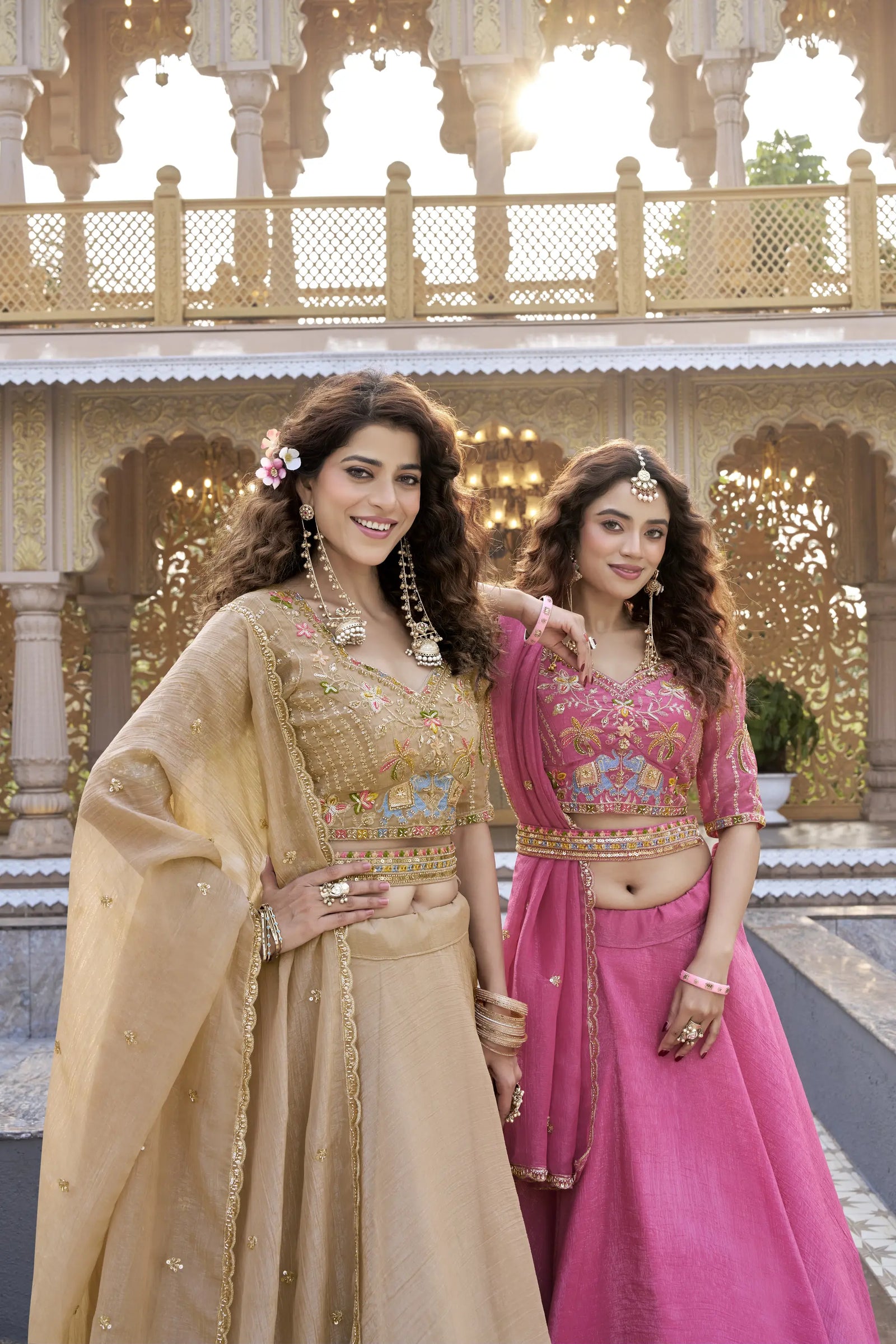 Two women in traditional Indian attire standing in front of an ornate architectural background.