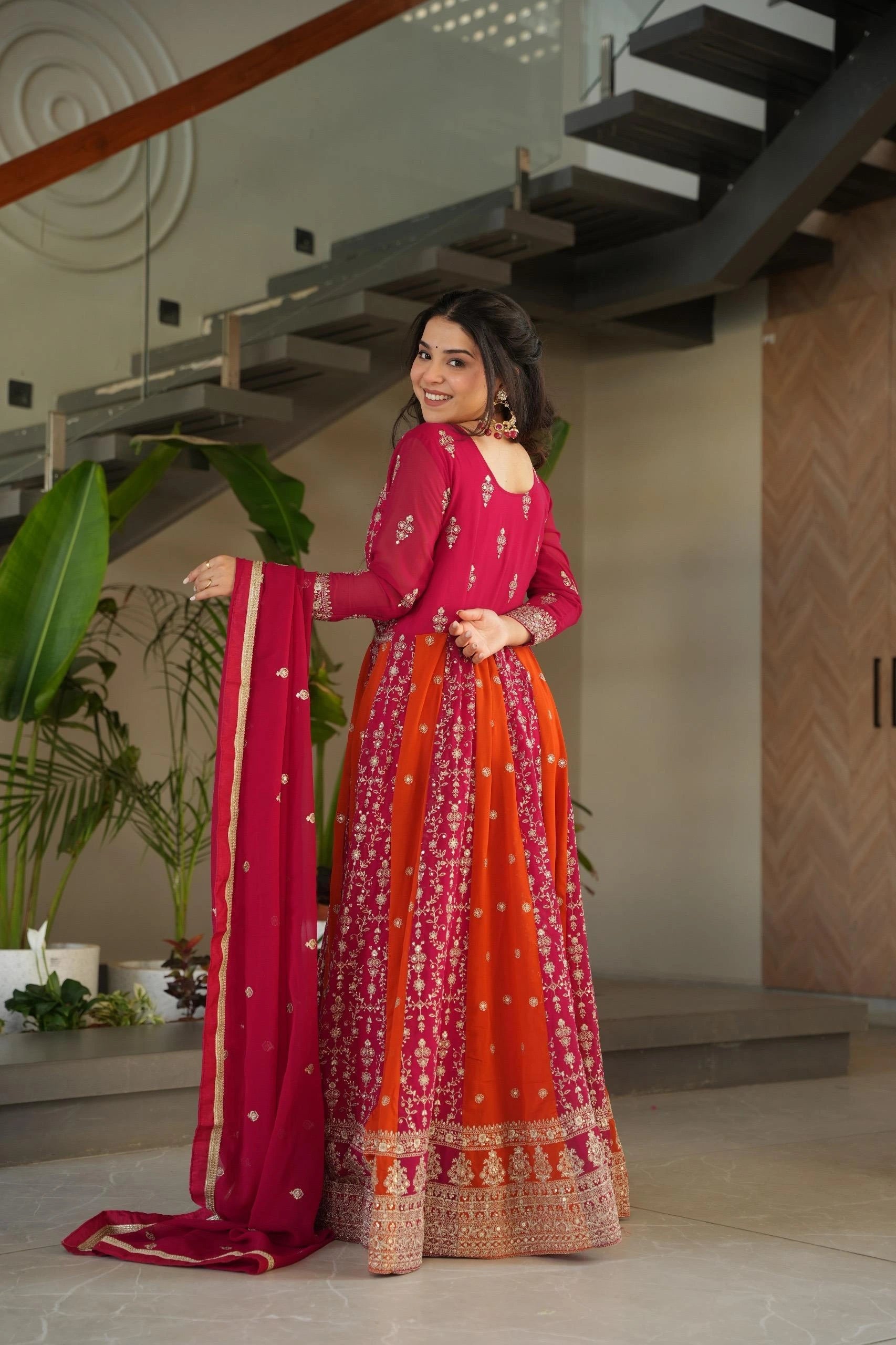 Woman in a red and orange traditional outfit standing indoors with plants and a staircase in the background.