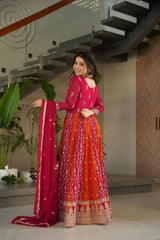 Woman in a red and orange traditional outfit standing indoors with plants and a staircase in the background.