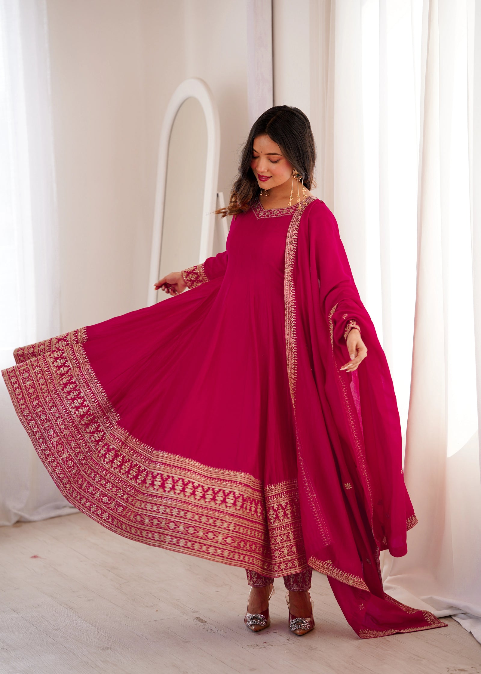 Woman in a vibrant pink traditional outfit with gold patterns, standing against a white curtain.
