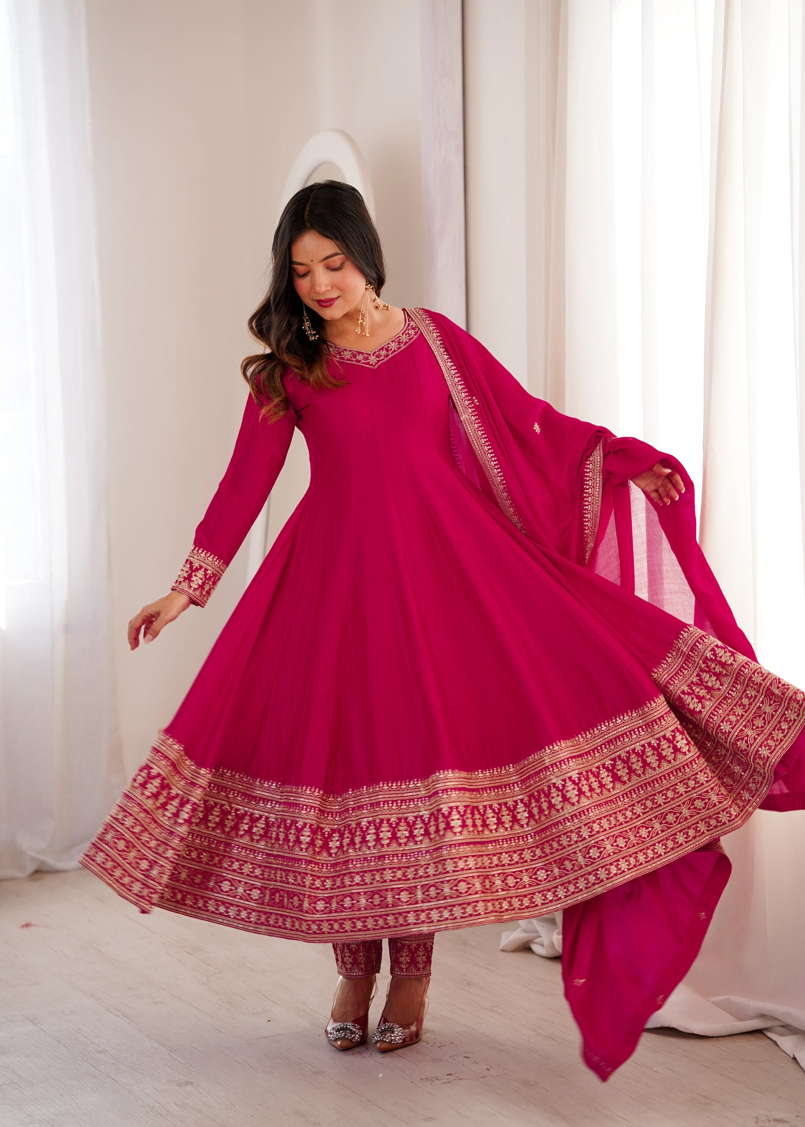 Woman wearing a pink traditional outfit with intricate patterns in a bright room.