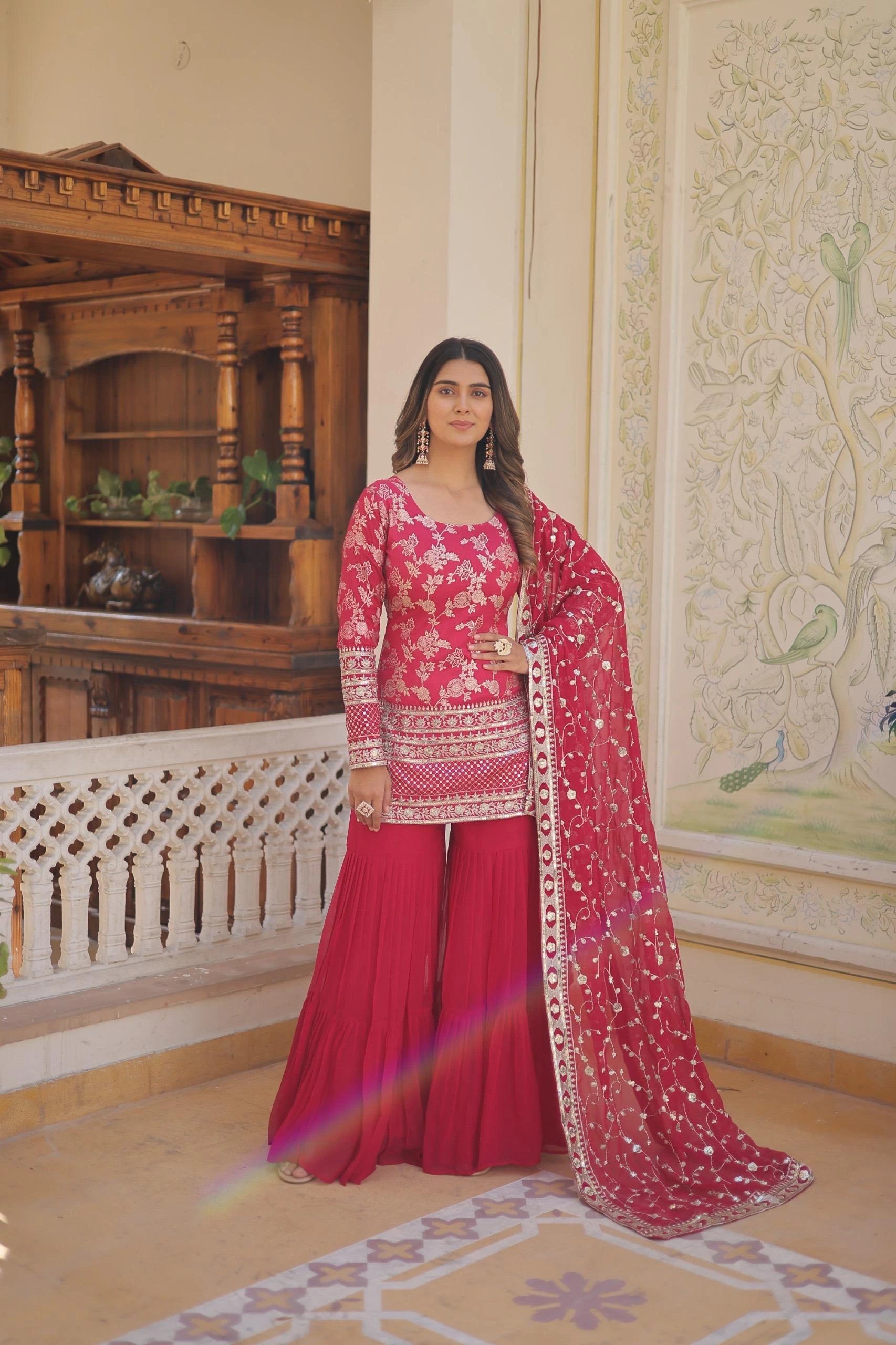 Woman in a traditional pink and red outfit standing in an ornate room.