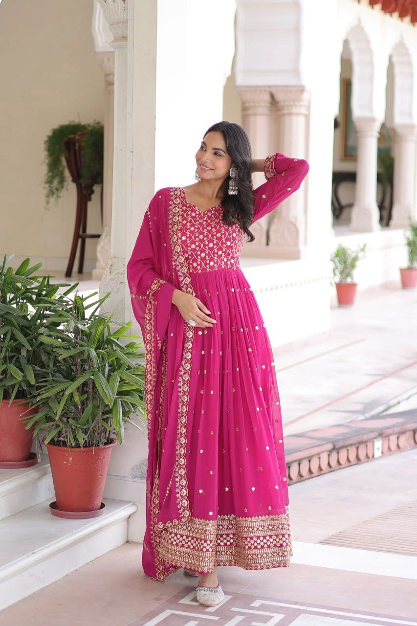 Woman in a pink traditional outfit standing in an outdoor setting with plants and architectural elements.