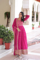 Woman in a pink traditional outfit standing in an outdoor setting with plants and architectural elements.