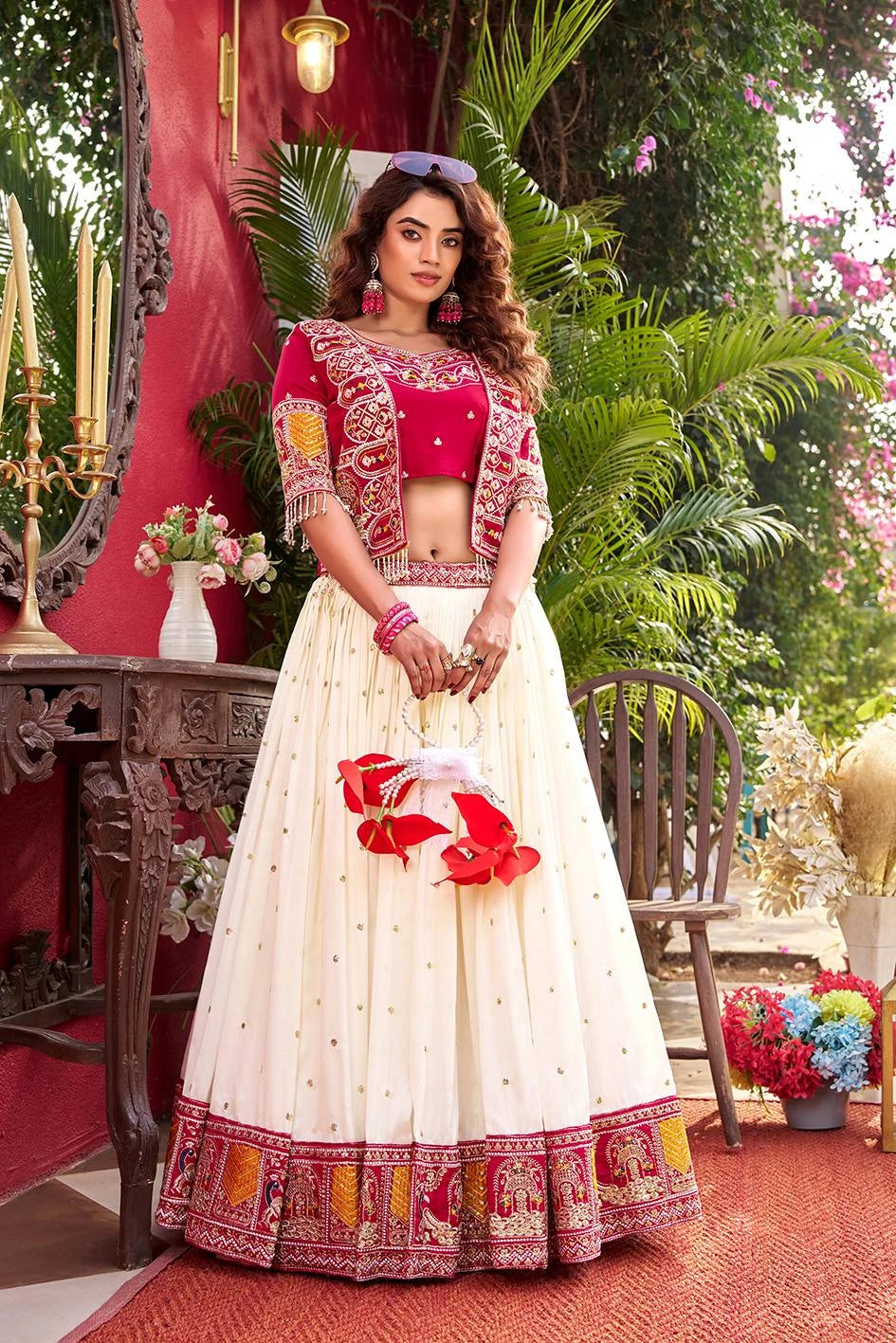 Woman in traditional outfit with red blouse and white skirt holding flowers, outdoor setting with greenery.