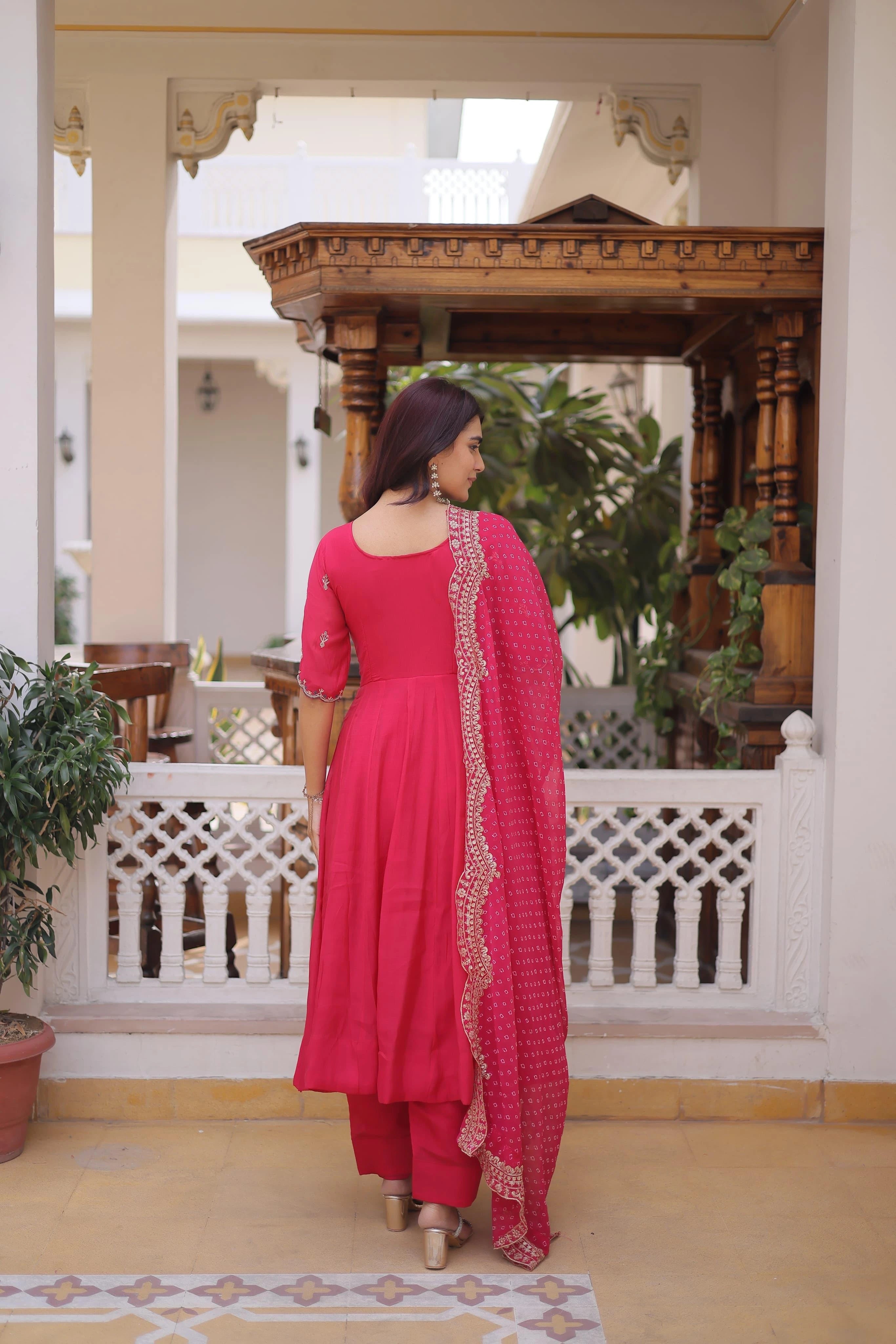 Woman in a pink traditional outfit standing in a decorative indoor setting with plants and wooden architecture.