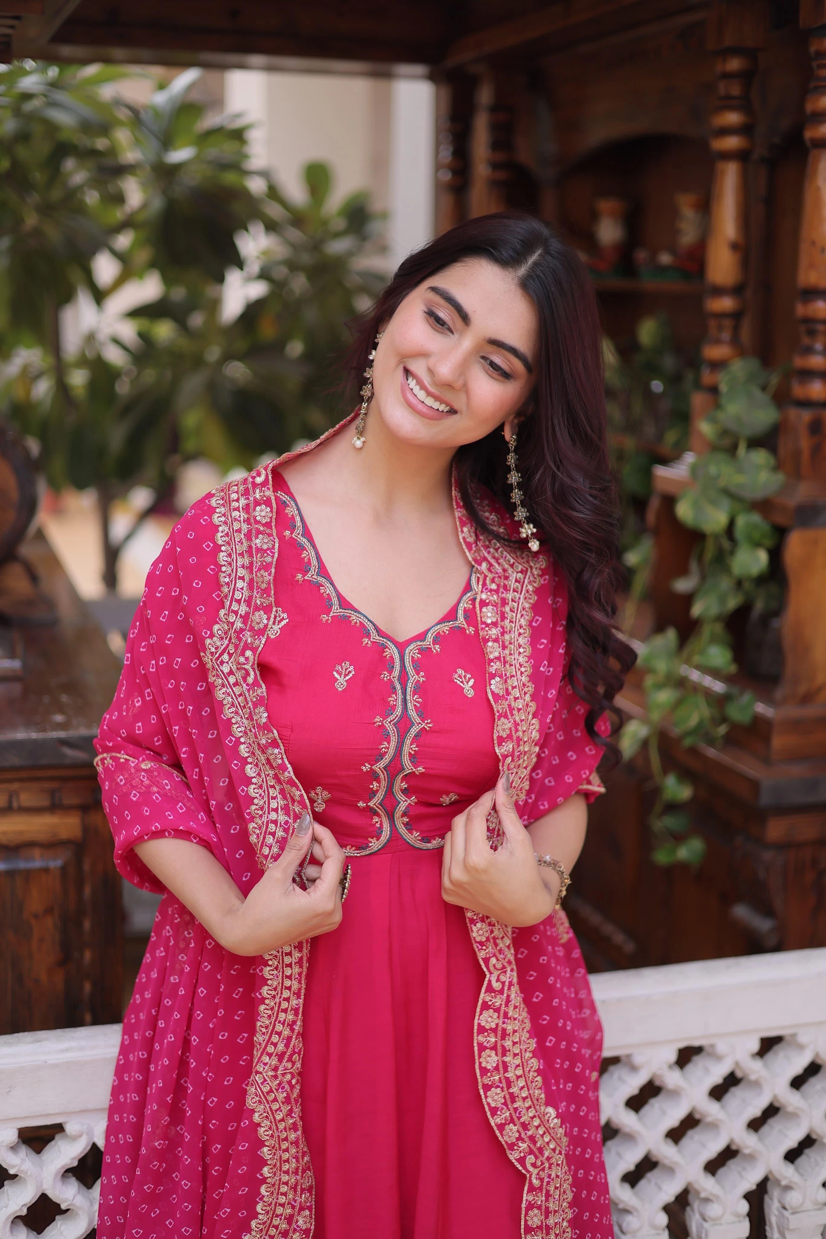Woman in a pink traditional outfit with gold details, standing outdoors with plants and wooden furniture in the background.