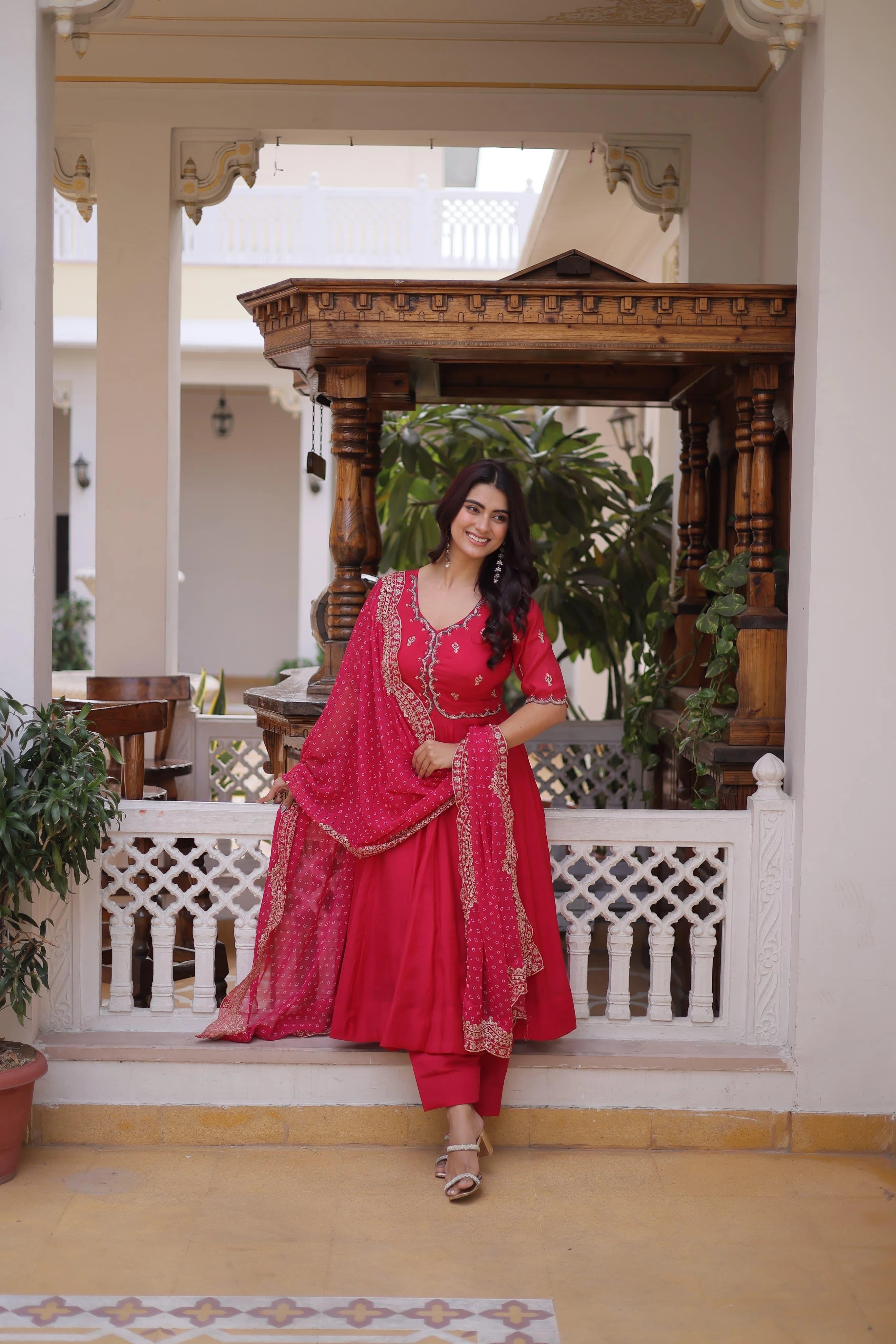 Woman in a red traditional outfit standing in an elegant indoor setting with plants and architectural elements.