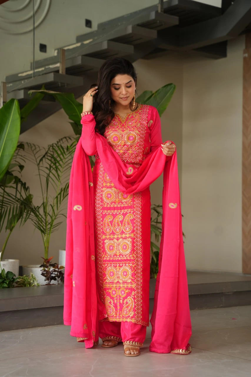Woman in a traditional pink and red embroidered outfit with a dupatta, standing indoors.