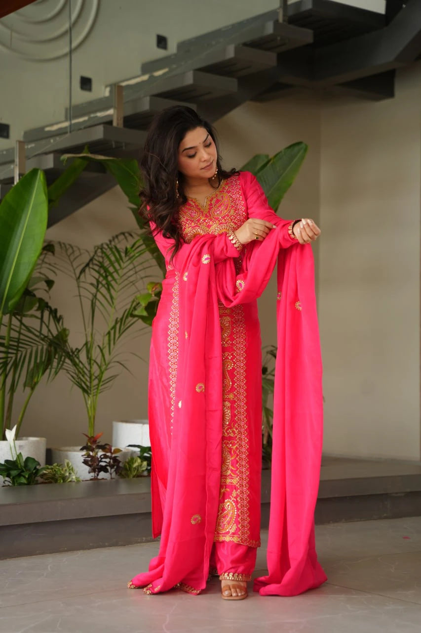 Woman in a bright pink traditional outfit with gold embroidery, standing indoors with plants in the background.