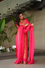 Woman in a bright pink traditional outfit with gold embroidery, standing indoors with plants in the background.