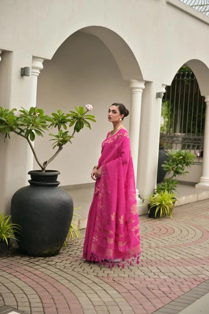 Woman in a pink saree standing in an outdoor setting with plants and arches.