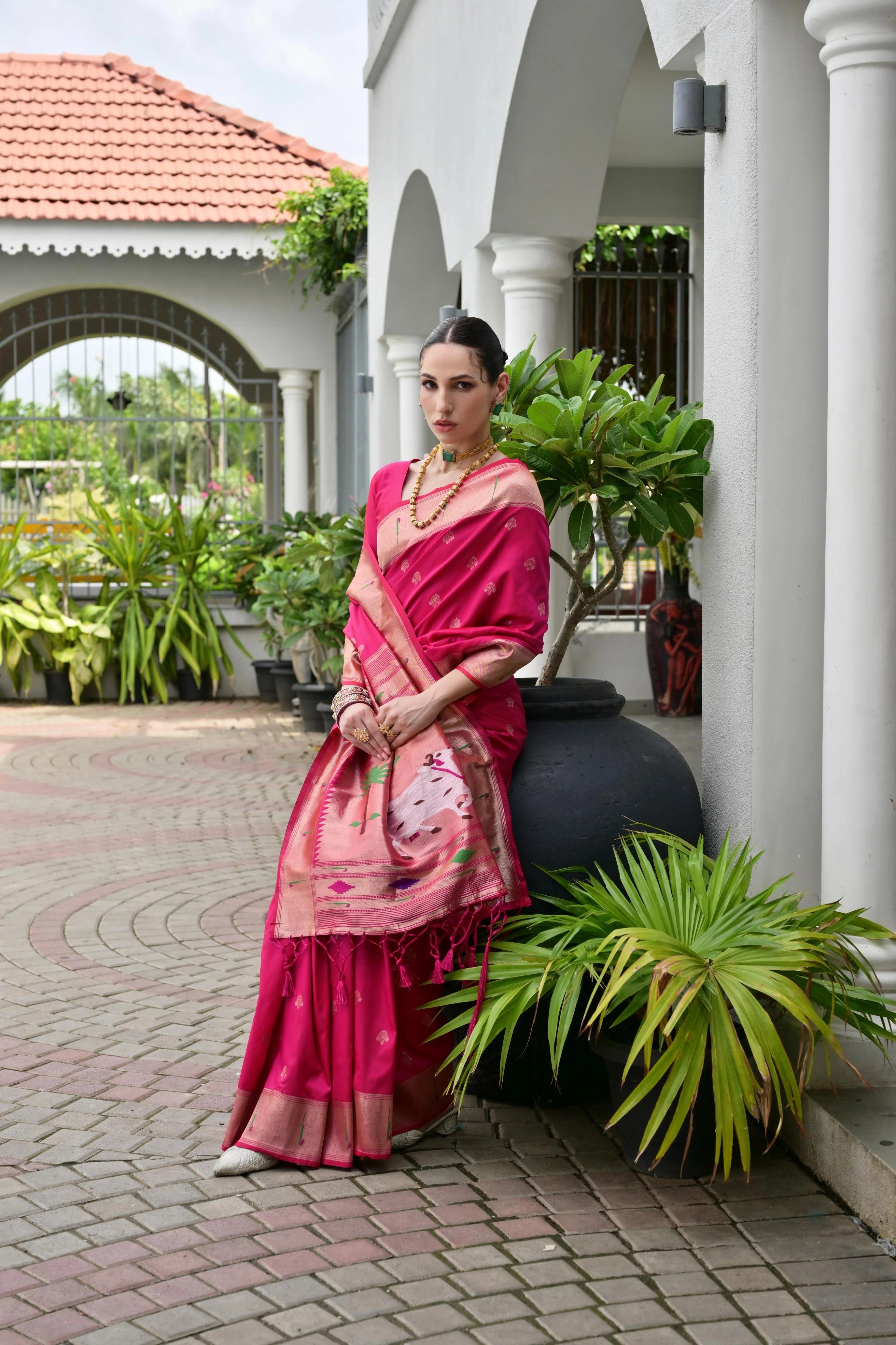 Woman in a pink saree standing outdoors near a building with plants.