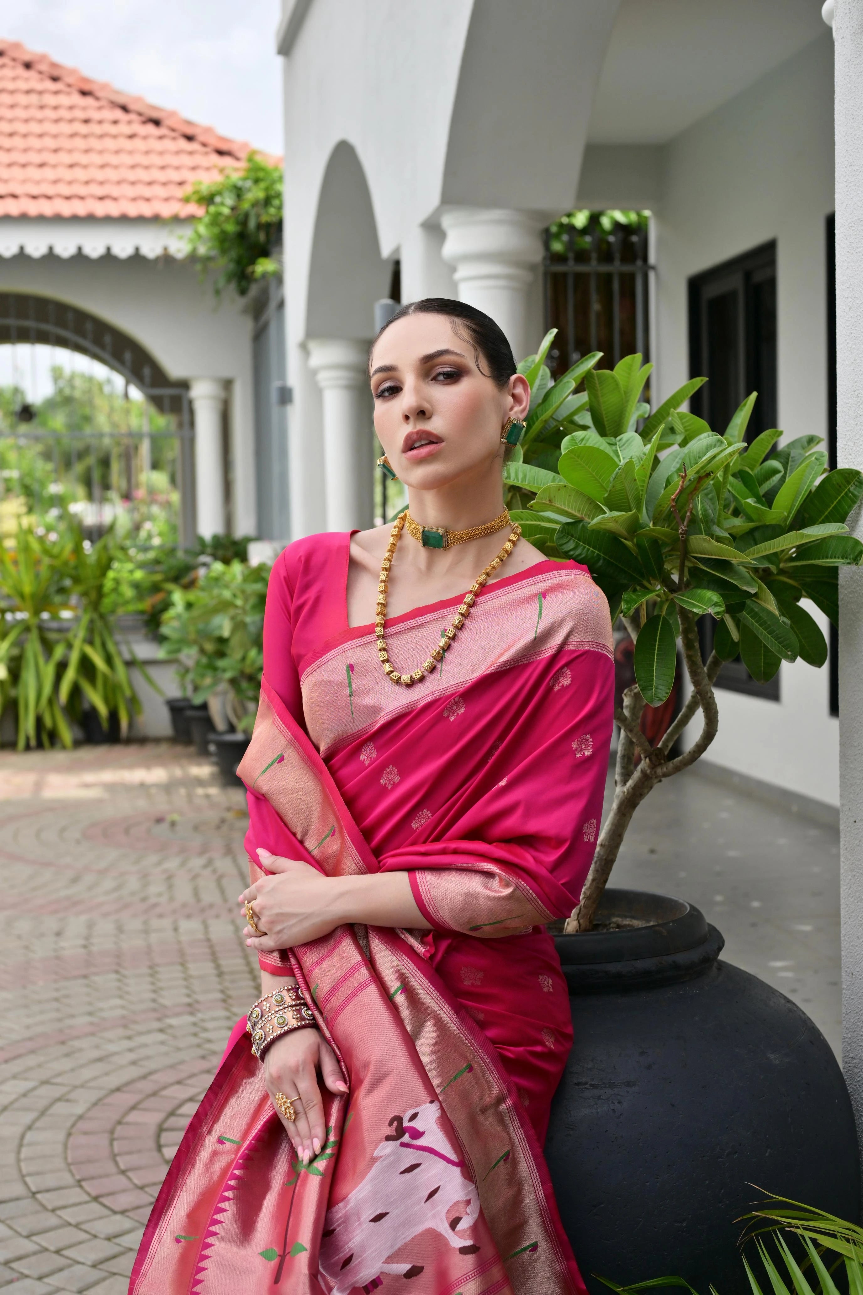 Woman in a pink saree standing outdoors with greenery and a building in the background
