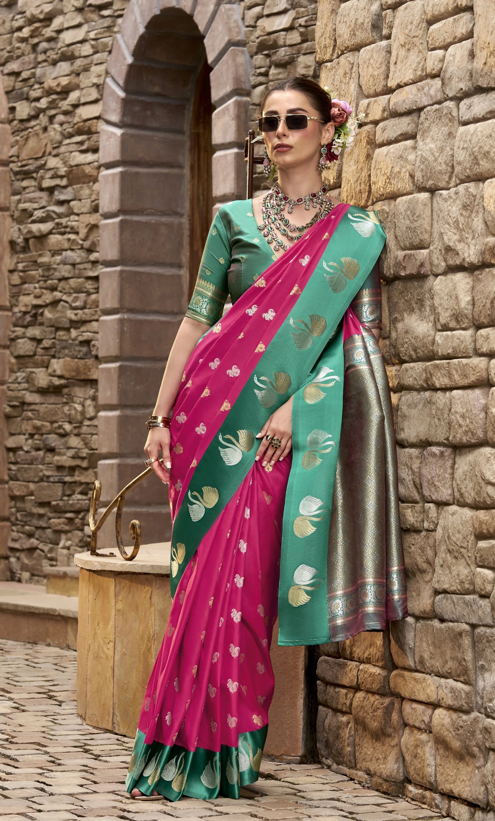 Woman in a pink and green saree standing against a stone wall.