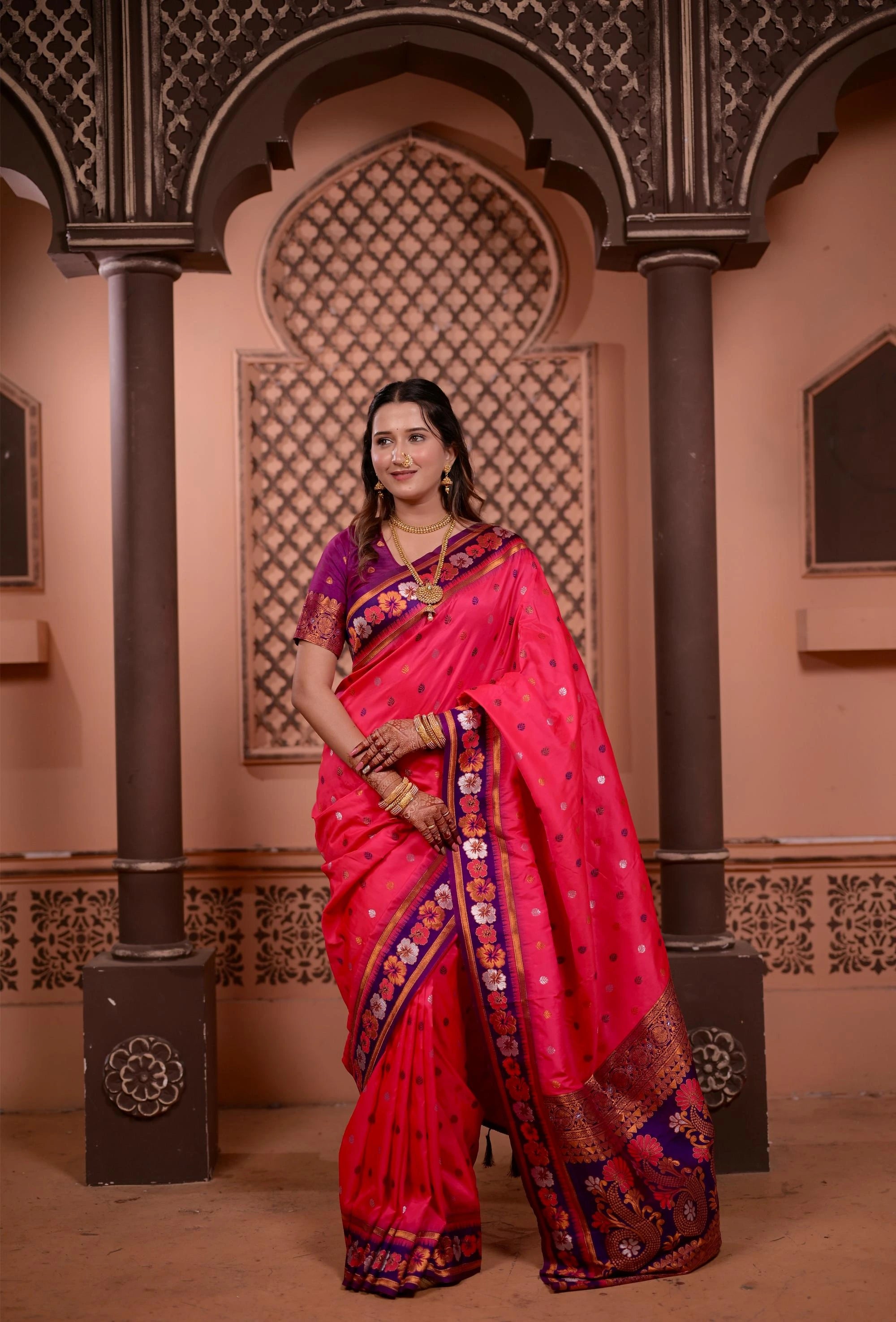 Woman in a vibrant pink saree with intricate patterns standing in an ornate architectural setting.