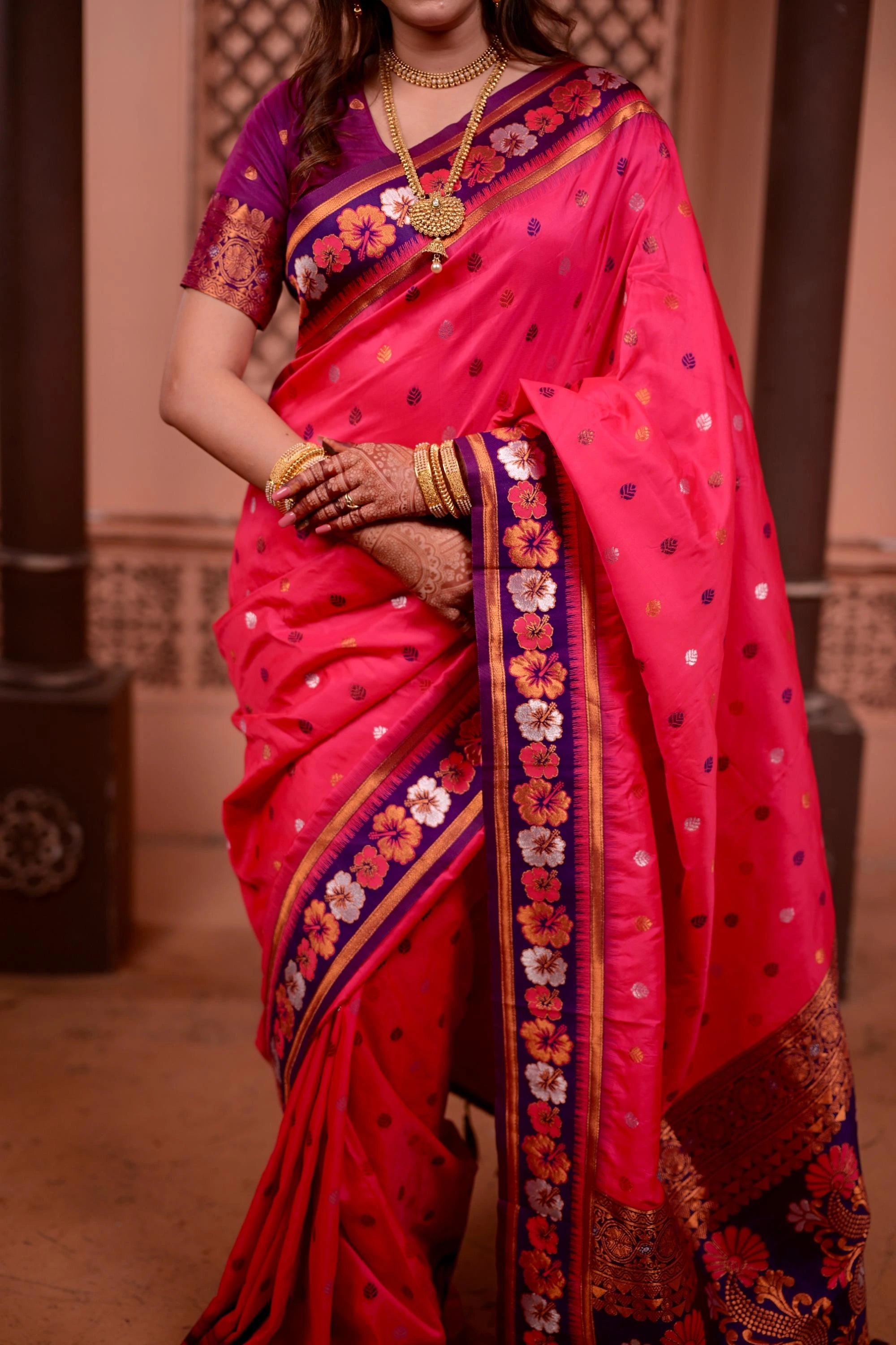 Woman wearing a vibrant pink saree with intricate designs, standing against a neutral background.