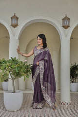 Woman in a purple saree standing in an outdoor setting with plants and arches.