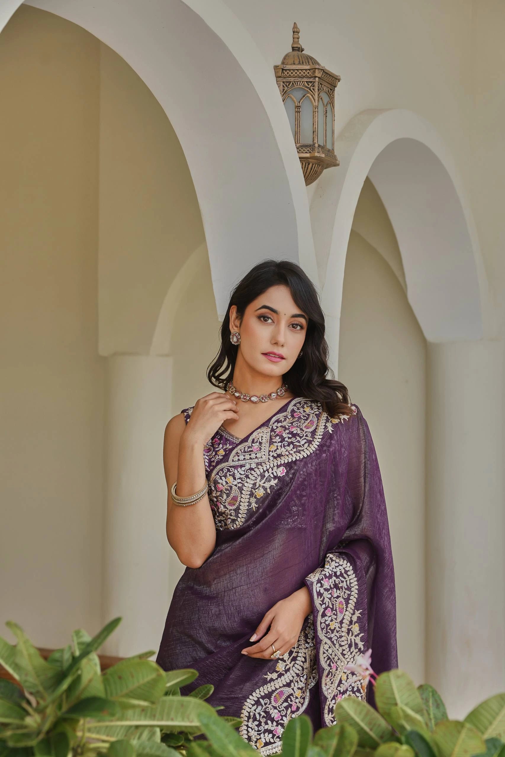 Woman in a purple saree standing in front of an archway with a lantern.
