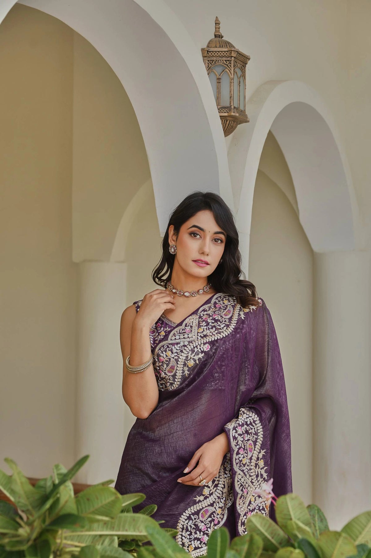 Woman in a purple saree standing in front of an archway with a lantern.