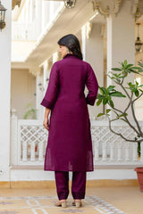 Woman in a purple outfit standing on a porch with white columns and a plant.