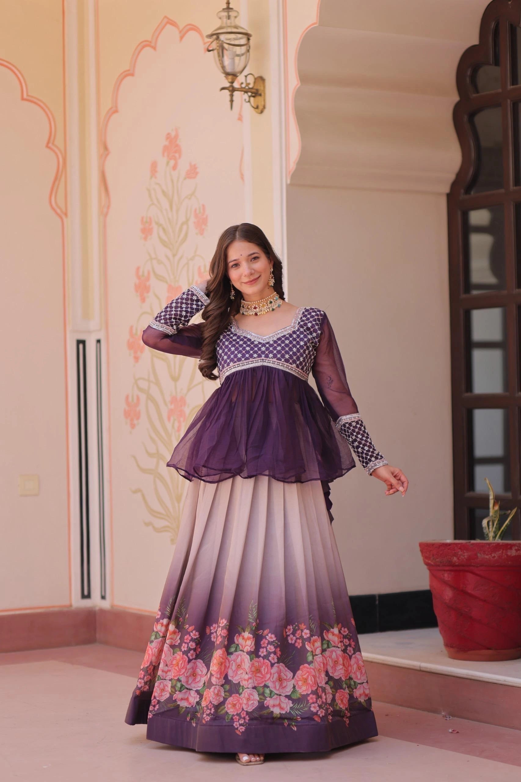 Woman in a traditional outfit with a purple top and floral skirt standing in a decorated room.