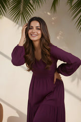 Woman in a purple dress standing against a beige wall with green leaves.
