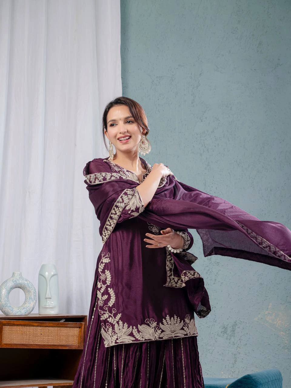 Woman in a purple traditional outfit with gold embroidery against a light blue wall.