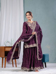 Woman in a traditional purple outfit with gold embroidery standing in a room with light blue walls and white curtains.